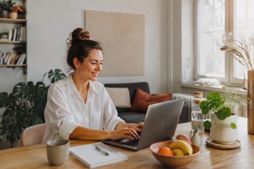 Eine Frau arbeitet zu Hause am Esstisch an ihrem Laptop, daneben Notizbuch, Tasse und eine Schale Obst.