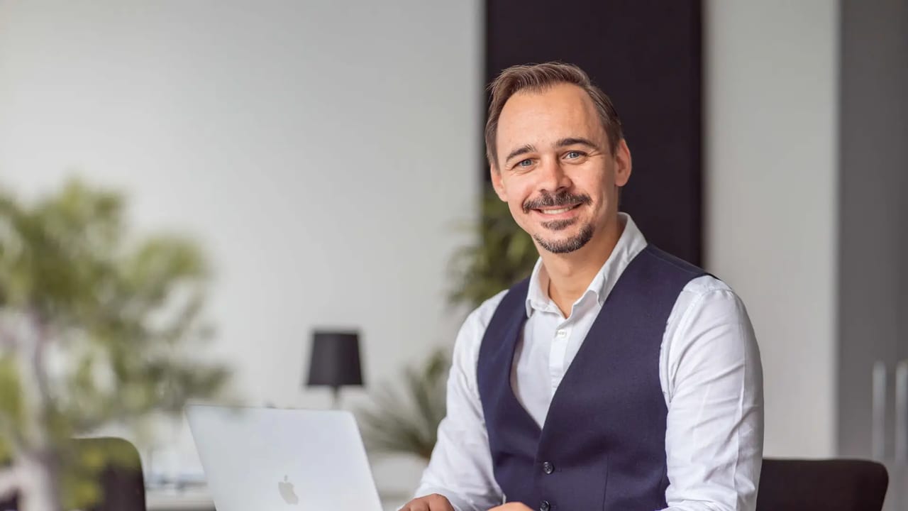 Alen Grajcevci am Schreibtisch im Enevo-Büro in Freising, heller Raum mit Pflanzen und aufgeklapptem Notebook.