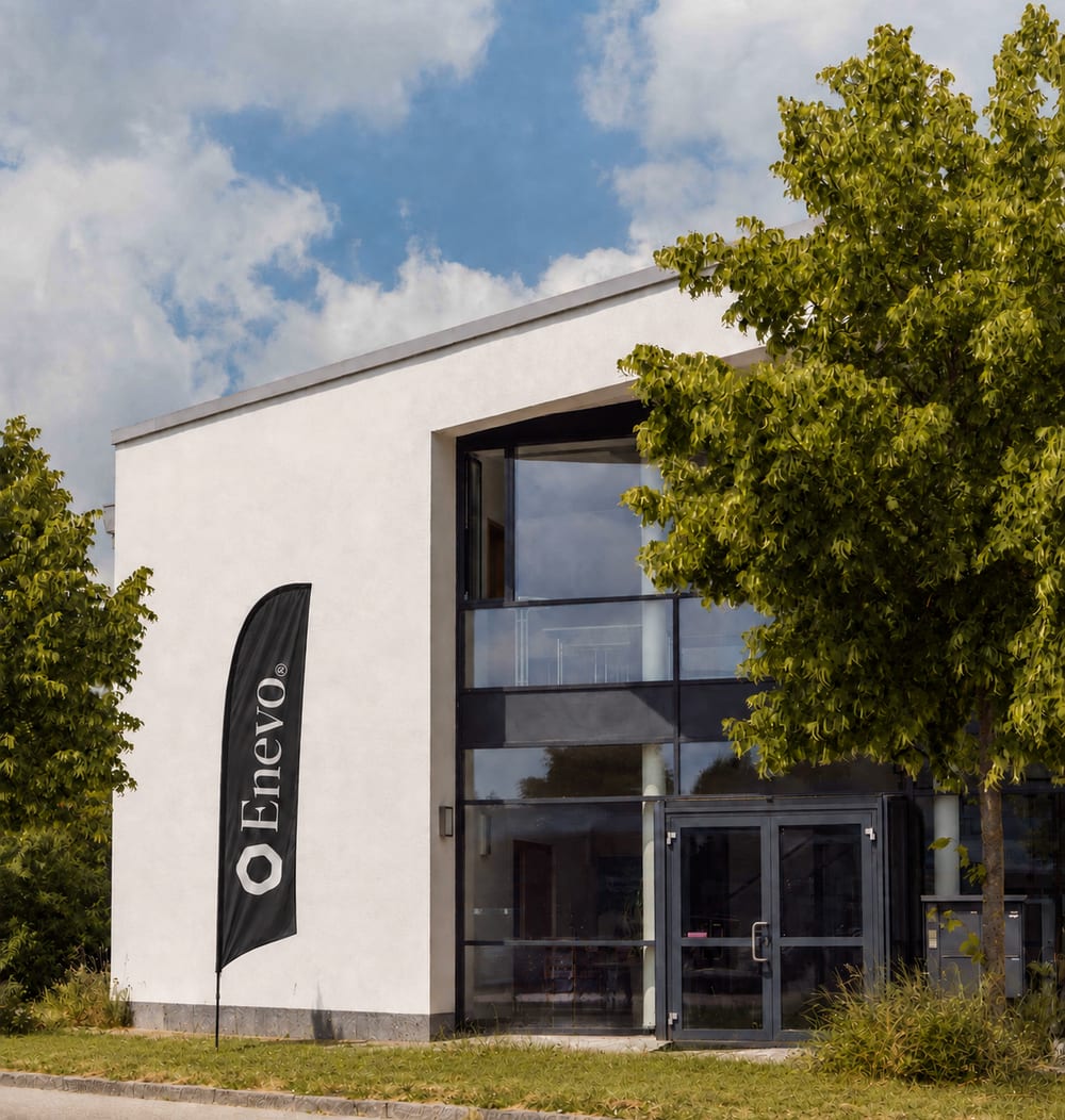 Bürogebäude von Enevo in Freising: helle Fassade mit ansteigendem Pultdach, große Glasfront am Eingang und davor eine Beachflag mit dem Enevo-Zeichen.