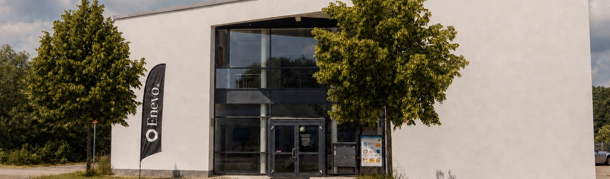 Bürogebäude von Enevo in Freising, davor zwei Bäume und eine Beachflag mit dem Enevo-Zeichen.