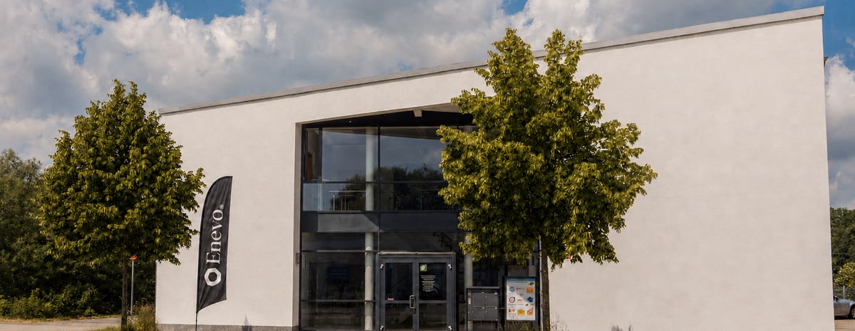 Das Firmengebäude von Enevo in Freising, davor zwei Bäume und eine Beachflag mit dem Enevo-Zeichen.
