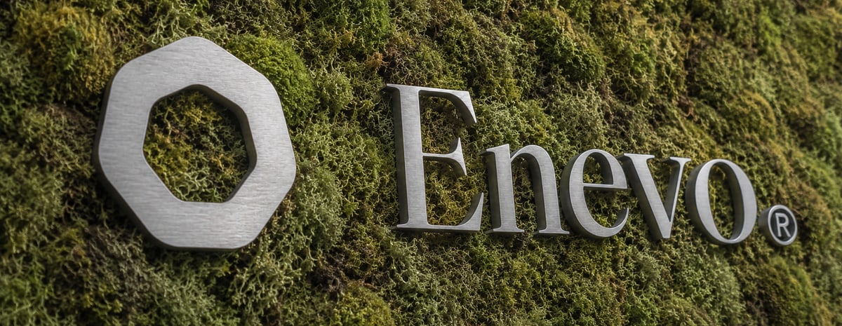 Das Enevo-Zeichen und der Schriftzug in gebürstetem Metall auf einer Wand aus Moos.